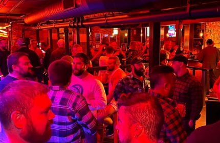 Chicago's Jackhammer gay bar party scene. A lively crowd of men in a dimly lit, industrial-chic space. Red and blue lights add to the ambiance.