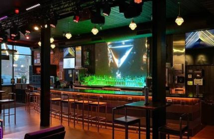 Interior of Jackhammer gay bar in Chicago. Bar stools line the counter, illuminated by colorful lights, creating a party atmosphere.