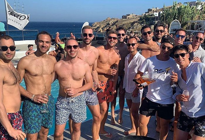 Gay group at Jackie O' Beach Club, Super Paradise, Mykonos. Men in swim trunks, some shirtless, holding drinks, enjoying the party.