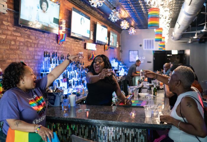 Inside Jeffery Pub, a lively gay bar in Chicago. People are toasting with shots at the bar, rainbow decorations visible. Gay travel.