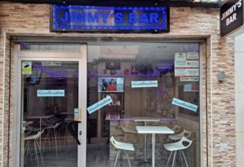 Jimmy's Bar, a gay bar in Benidorm. The entrance is visible, with tables and chairs inside. A blue LED sign displays the bar's name.