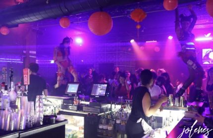Jolene's Queer Bar in San Francisco: Performers dance above the bar in a crowded, purple-lit space with orange lanterns. Gay nightlife.