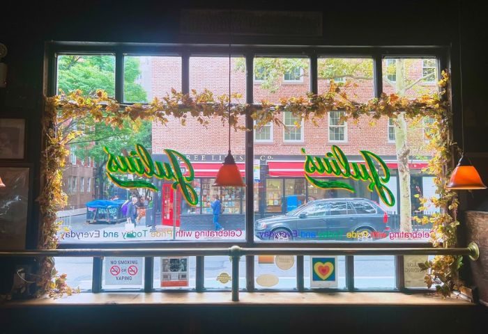 Julius' Gay Bar in New York City. View through the window with the bar's name in green neon. A car and people are visible outside.