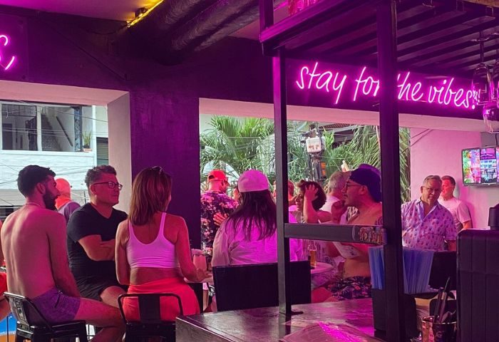 Kevin's Hideaway gay bar in Puerto Vallarta. People at the bar, neon sign says Stay for the vibes. Gay travel, nightlife.