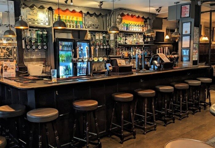 The Kings Arms Soho bar interior. London's best gay bear bar near a gay sauna. Bar stools line the counter with rainbow flag decor above.