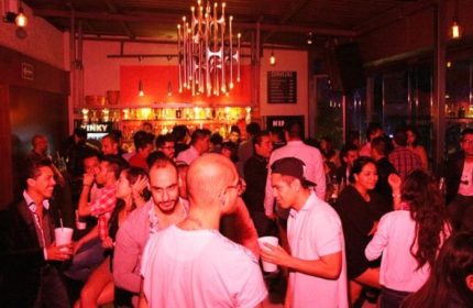 Inside Kinky Bar, a gay bar in Mexico, patrons enjoy drinks. The scene is lively with people socializing under warm, red lighting.