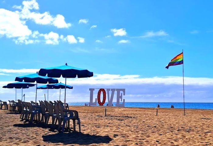 Gran Canaria gay beach scene: Rainbow flag, LOVE sign, beach umbrellas, and chairs on the sand. Gay bar kiosk in the background.