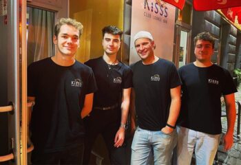 Four men in front of Kisss Bar Vienna, a gay club-lounge-bar. They're wearing branded t-shirts. Gay travel advice in Vienna.
