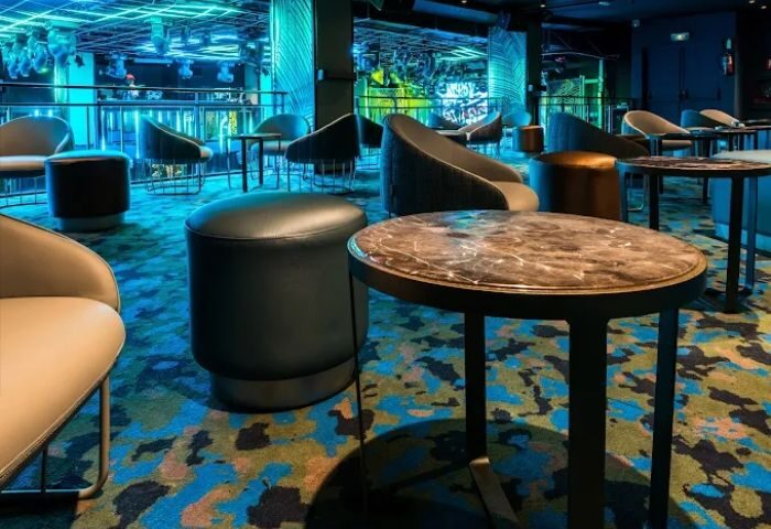 Kluster Rebel Gay Club in Madrid lounge area. Tables, chairs, and ottomans on a patterned carpet. Blue lighting. Gay travel advice.