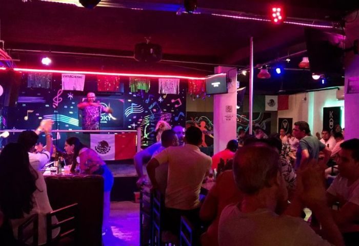 Kooky Karaoke in Puerto Vallarta: Gay men enjoy a lively karaoke night at a bar with a DJ, stage, and Mexican flags. Nightlife scene.