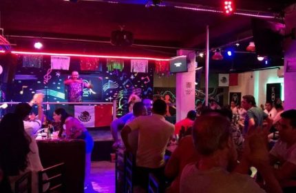 Kooky Karaoke in Puerto Vallarta: Gay men enjoy a lively karaoke night at a bar with a DJ, stage, and Mexican flags. Nightlife scene.