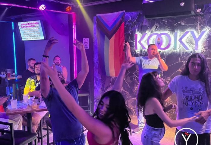 Gay men enjoy Kooky Karaoke in Puerto Vallarta. People dance as a man sings karaoke in front of a Progress Pride flag.