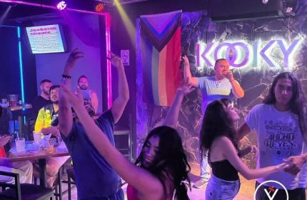 Gay men enjoy Kooky Karaoke in Puerto Vallarta. People dance as a man sings karaoke in front of a Progress Pride flag.