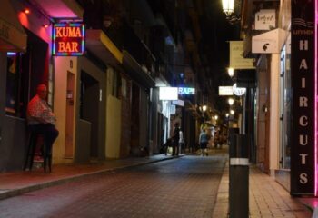 Night view of Kuma Bar, Benidorm's gay bar. Man sits outside. Street scene with neon signs, a party spot for gay men's travel.