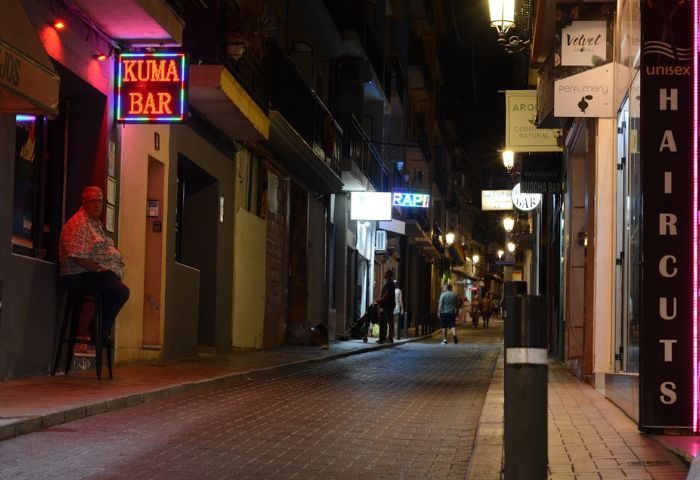 Night view of Kuma Bar, Benidorm's gay bar. Man sits outside. Street scene with neon signs, a party spot for gay men's travel.