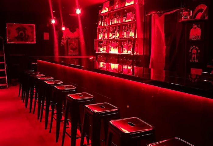 Interior of Kuma Bar, a gay bar in Benidorm. Red lighting, bar stools, shelves of liquor bottles. Nightlife and party scene.