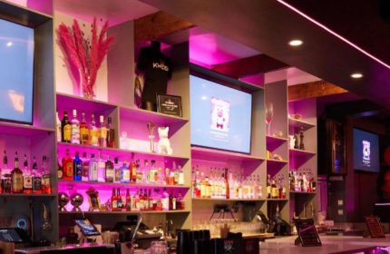 Kween Cocktails Kitchen bar in Montreal. Shelves of liquor bottles, TVs, pink lighting. Gay travel advice for Montreal's nightlife.