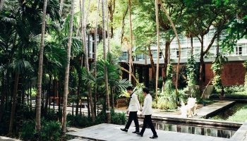 Pullman Bangkok King Power LGBTQ wedding. Gay couple in white suits walking hand in hand in a tropical garden at the hotel.