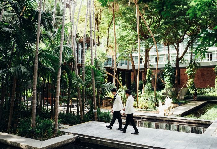 Pullman Bangkok King Power LGBTQ wedding. Gay couple in white suits walking hand in hand in a tropical garden at the hotel.