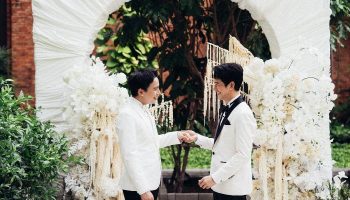 Gay wedding at Pullman Bangkok King Power. Two grooms in white suits holding hands under a floral arch. LGBTQ+ travel in Thailand.