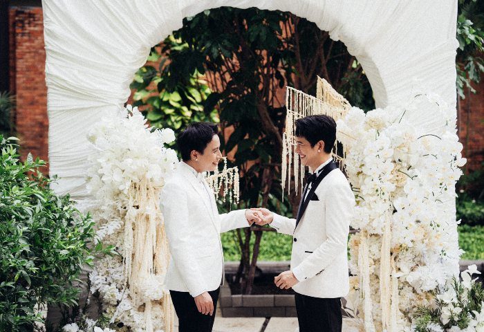 Gay wedding at Pullman Bangkok King Power. Two grooms in white suits holding hands under a floral arch. LGBTQ+ travel in Thailand.