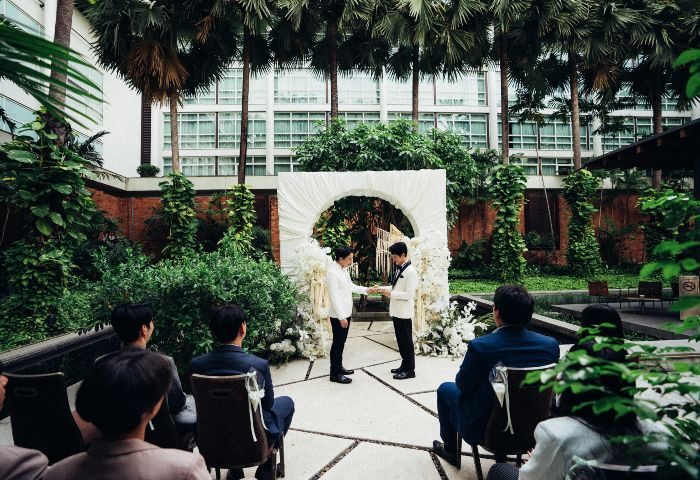 Gay wedding at Pullman Bangkok King Power. Two grooms exchange rings under a floral arch, surrounded by guests in a lush garden setting. LGBTQ+ travel.