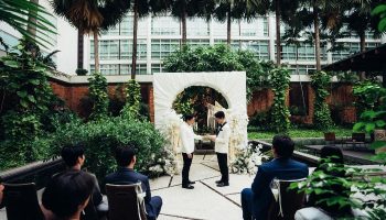 Gay wedding at Pullman Bangkok King Power. Two grooms exchange rings under a floral arch, surrounded by guests in a lush garden setting. LGBTQ+ travel.