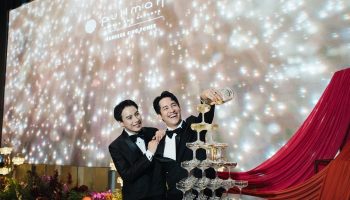 Gay wedding at Pullman Bangkok King Power. Two grooms in tuxedos pour champagne into a tower of glasses. LGBTQ+ wedding celebration.
