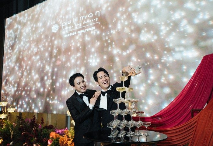 Gay wedding at Pullman Bangkok King Power. Two grooms in tuxedos pour champagne into a tower of glasses. LGBTQ+ wedding celebration.