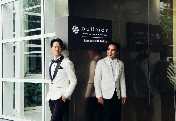Gay wedding at Pullman Bangkok King Power. Two grooms in white tuxedos smile. LGBTQ wedding in Thailand. Gay travel advice.