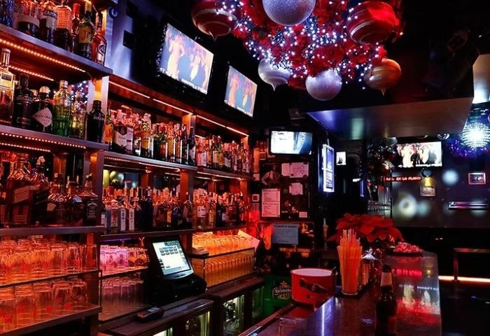 Interior of LL Bar, a gay bar in Madrid. Shelves of liquor bottles, TVs, and festive decorations create a vibrant nightlife scene.