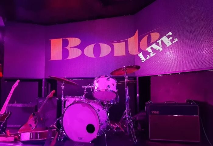Stage view at La Boite Gay Club, Madrid. Drum set and guitars under purple lights. Boite Live sign. Gay nightlife, travel advice.