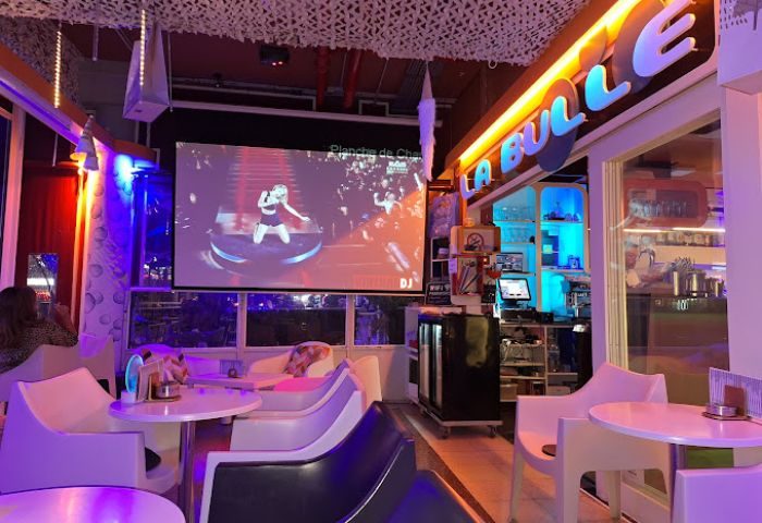 Interior of La Bulle Gay Bar in Gran Canaria. Screen showing a singer, modern furniture, and colorful lighting create a vibrant atmosphere.