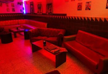 Interior of La Casa de la Pradera, a gay bar in Barcelona, featuring red lighting, sofas, tables, and posters on the wall.