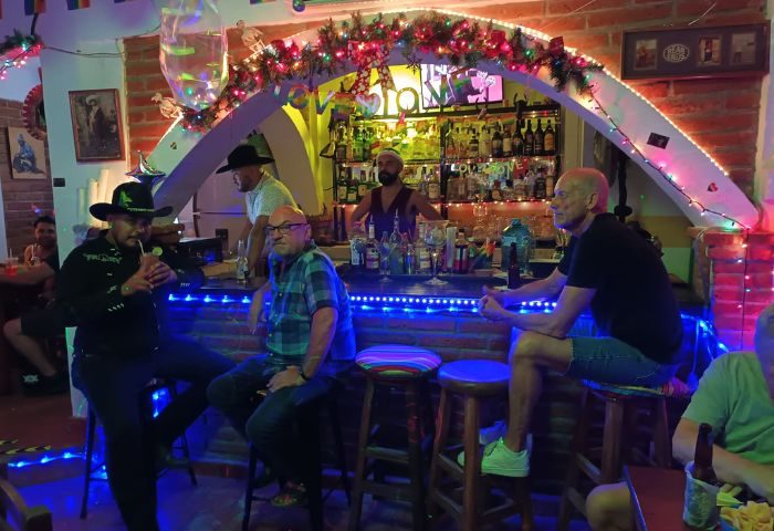 La Cueva Cantina, a gay bar in Puerto Vallarta. Men at the bar, bartender, festive lights, and rainbow flags create a welcoming atmosphere.