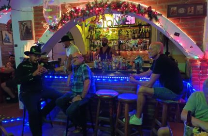 La Cueva Cantina, a gay bar in Puerto Vallarta. Men at the bar, bartender, festive lights, and rainbow flags create a welcoming atmosphere.