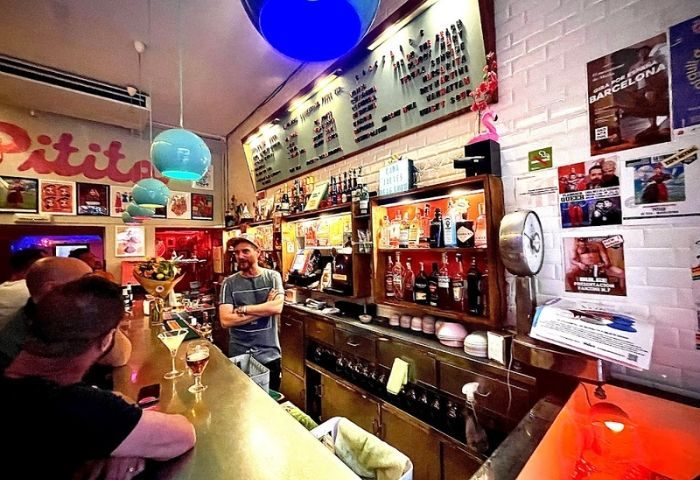 Inside La Federica Gay Bar in Barcelona. Bartender stands behind the bar, drinks served, and posters adorn the walls. Gay travel advice.