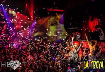 La Troya gay club in Ibiza's HEART, a vibrant party scene. Confetti rains down on the crowd, stage lights illuminate a drag queen performer.