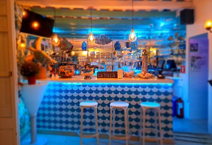 Interior of La Villa gay bar, Sitges. Bar stools line a blue-and-white scale-patterned counter. Bottles are displayed on shelves.