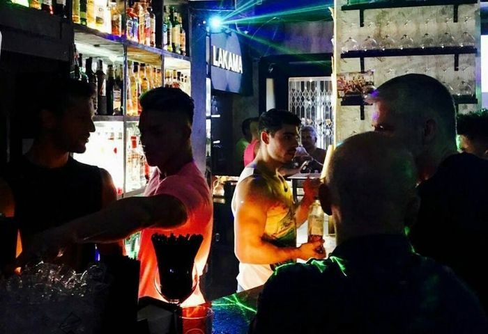 La Kama Gay Bar in Madrid: Men at the bar with drinks, shelves of liquor bottles, and laser lights. Gay nightlife in Madrid, Spain.