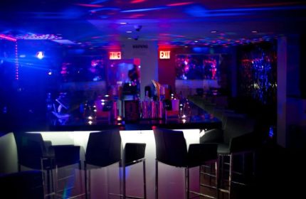 Lambda Lounge gay bar in NYC. Bar stools line the bar with a white front panel. Blue and red lights illuminate the ceiling.