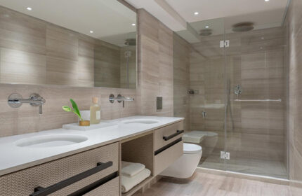 Dan Tel Aviv Hotel bathroom. Modern design with double sinks, light stone walls, and glass shower. Gay honeymoon travel inspiration.
