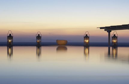 Porto Mykonos Hotel infinity pool at sunset. Gay-friendly hotel in Mykonos Town, Chora. Lanterns glow, reflecting in the still water.