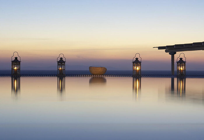 Porto Mykonos Hotel infinity pool at sunset. Gay-friendly hotel in Mykonos Town, Chora. Lanterns glow, reflecting in the still water.