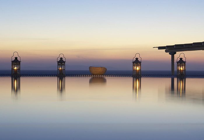 Porto Mykonos Hotel infinity pool at sunset. Gay-friendly hotel in Mykonos Town, Chora. Lanterns glow, reflecting in the still water.