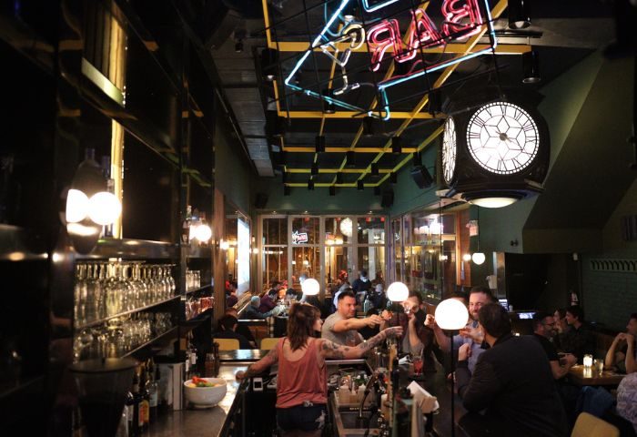Interior of Le Saloon Gay Bar in Montreal. Bartender serving patrons at the bar. Neon bar sign and clock hanging from the ceiling.