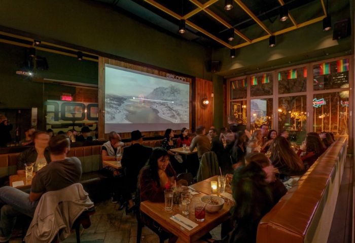 Le Saloon Gay Bar in Montreal: Interior view of a lively bar with patrons at tables, a large screen displaying a landscape, and rainbow flags.