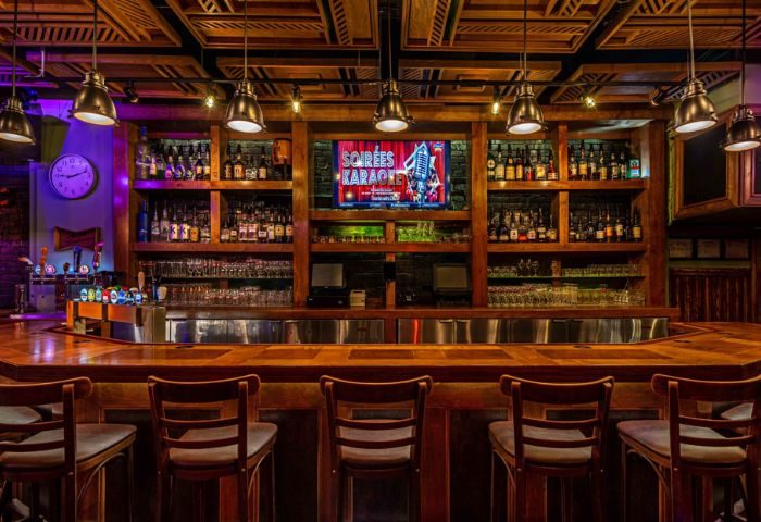 Le Taverne Normandie, a gay bar in Montreal. A warm interior with a wooden bar, stools, and shelves stocked with drinks. Karaoke screen visible.