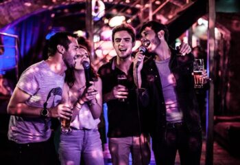 Le Clan Prague: Gay men singing karaoke, holding beers, and having fun in a nightclub with purple lighting. Gay travel advice.