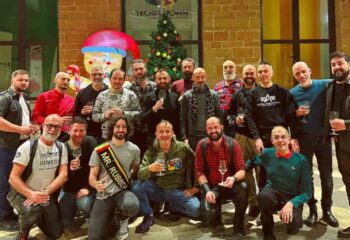 Leather Club Roma event: Group of gay men celebrating Christmas, some in leather attire, holding wine glasses, festive atmosphere.
